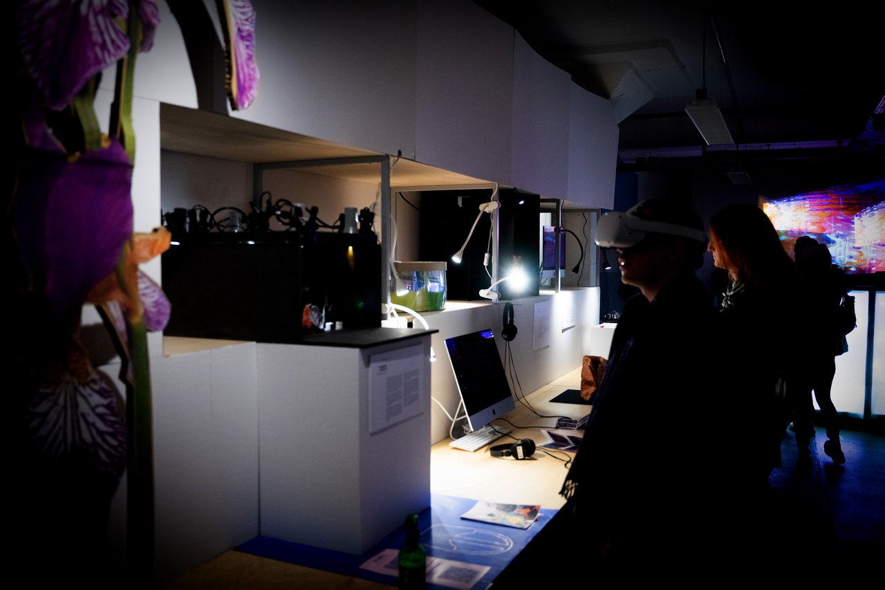 Innenaufnahme eines Technologie- oder Messebereichs mit Ausstellungsständen, auf denen elektronische Geräte und Bildschirme präsentiert werden, und Personen, die die Ausstellung besichtigen.