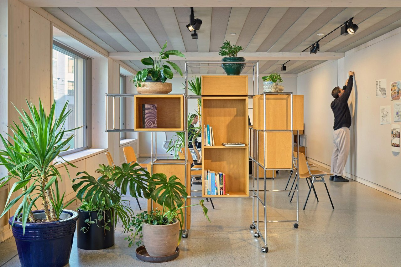 Modernes, helles Bürointerieur mit modularen Regalsystemen, Pflanzen und einem Mitarbeiter, der an einer Wand hängt.