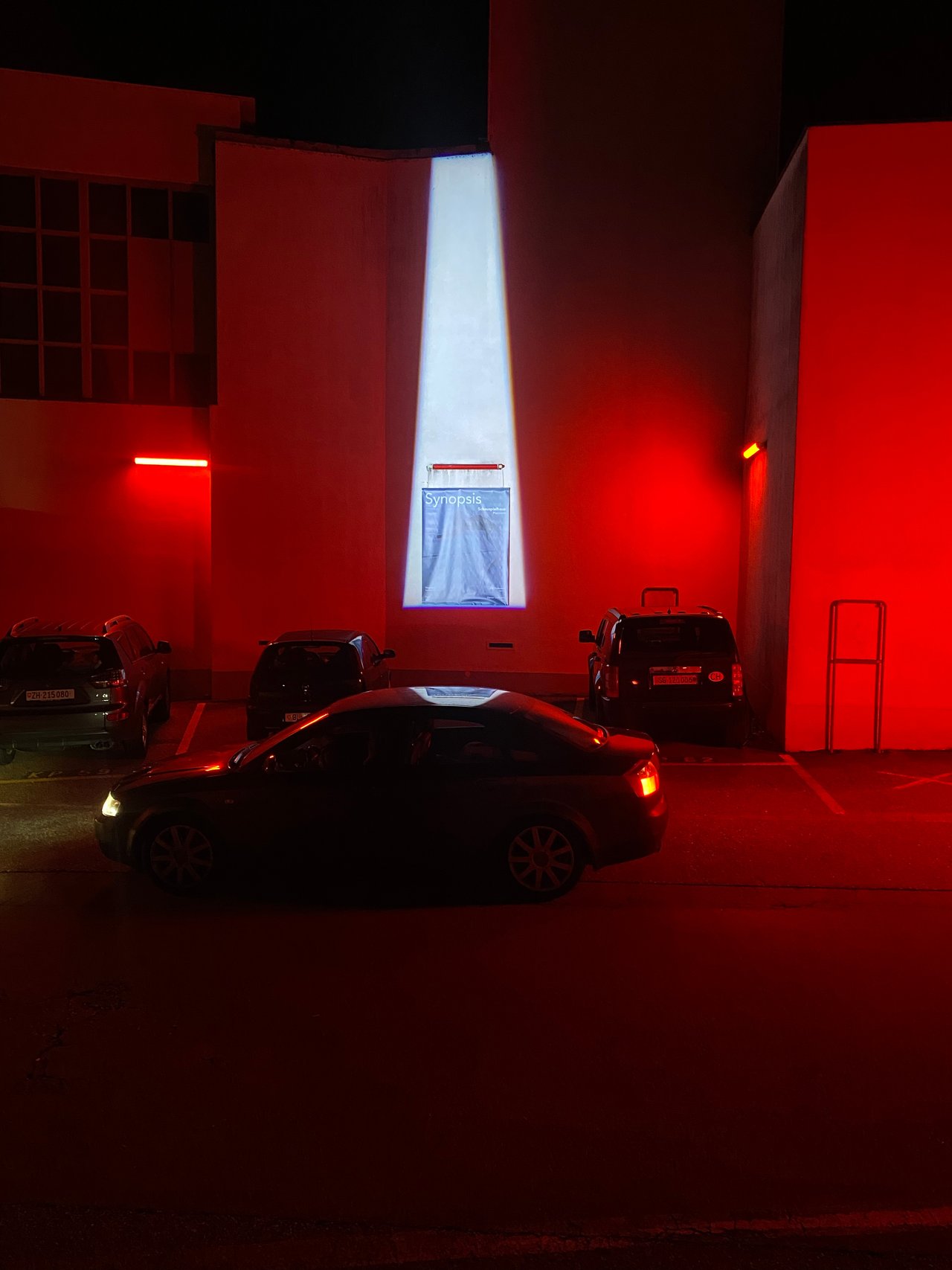 Abendaufnahme von mehreren Autos vor einer modernen, beleuchteten Fassade mit einem großen, vertikalen Fenster und rotem Akzentlicht